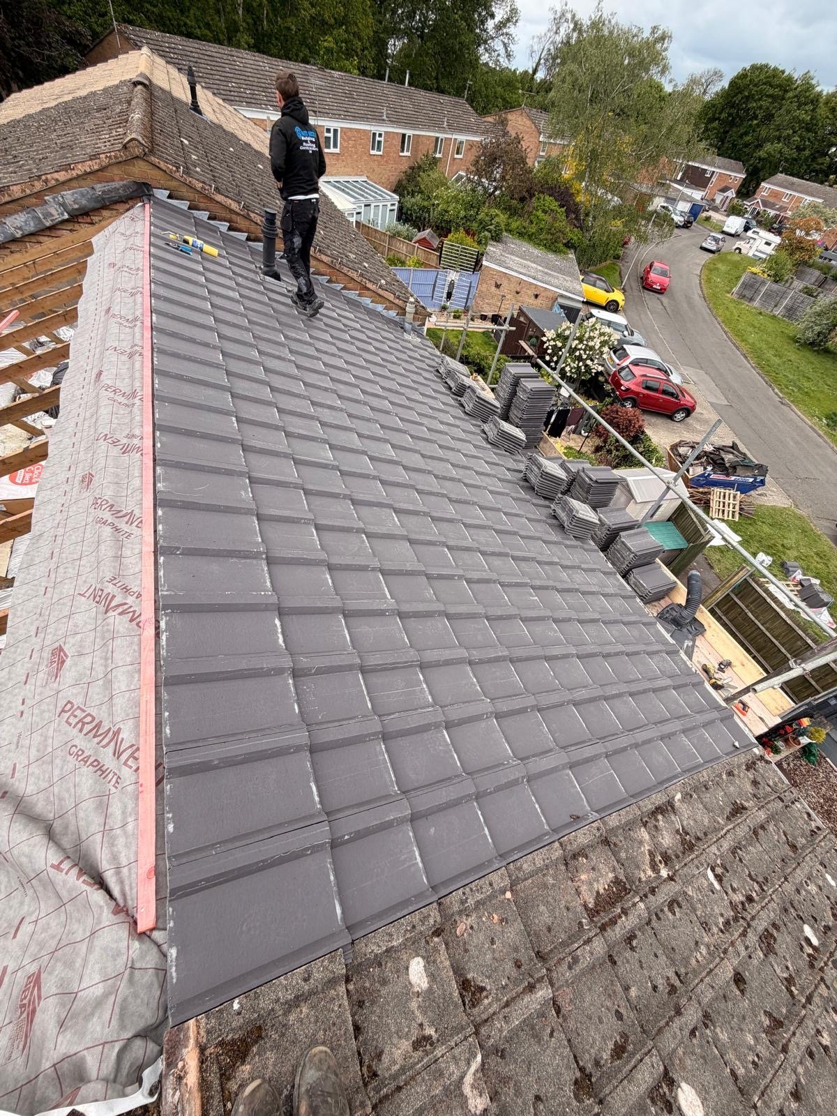 Roofer in a SkyMantle hoodie laying new grey interlocking tiles on a pitched roof, with fresh tile stacks alongside